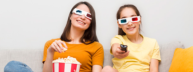 mujer y niña sentadas en sofá con gafas de cine, palomitas y control remoto en mano, viendo una película deportiva