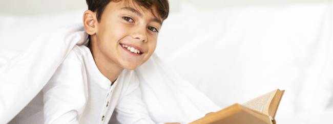 Niño sonriendo debajo de una sabana mientras lee un libro 